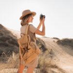 attractive-stylish-young-woman-khaki-dress-desert-traveling-africa-safari-wearing-hat-backpack-taking-photo-vintage-camera_285396-9174